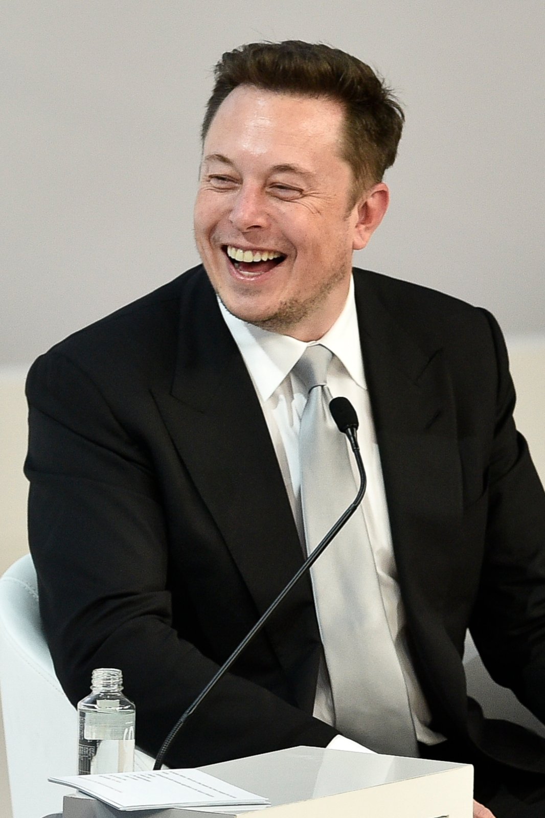 Elon Musk sitzt lachend vor einem Mikrofon