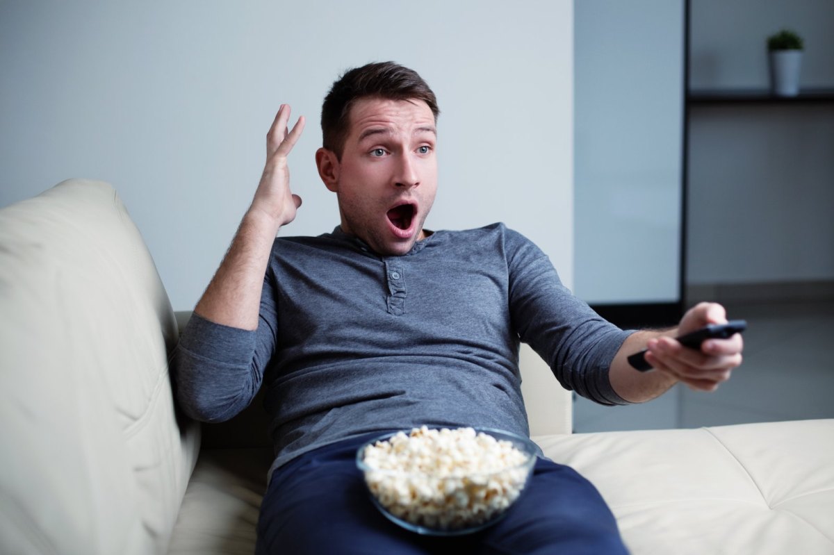 Ein Mann sitzt mit Popcorn erschrocken vor dem Fernseher.