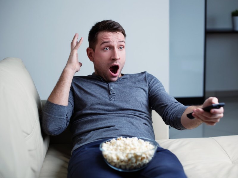 Ein Mann sitzt mit Popcorn erschrocken vor dem Fernseher.
