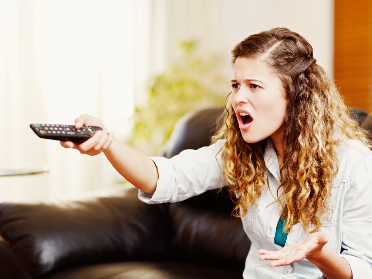 Frau zielt mit einer Fernbedienung ärgerlich auf einen Fernseher.