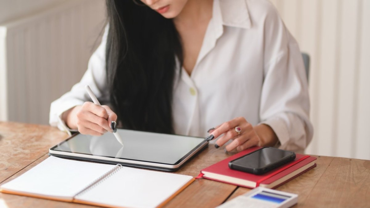 Eine Frau sitzt am Schreibtisch und schreibt mit einem Pen auf ihrem Tablet.