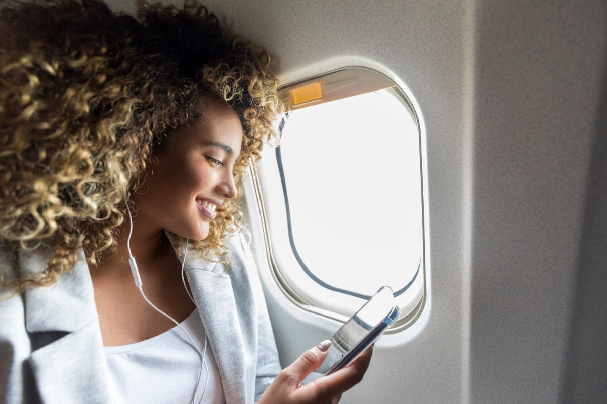 Frau sitzt mit Handy und Kopfhörern am Fenster im Flugzeug.