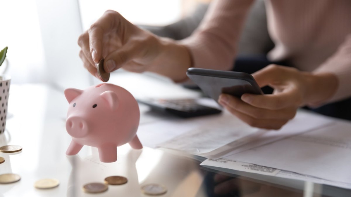 Frau hält Handy in der Hand und schmeißt Geld in ein Sparschweinchen.