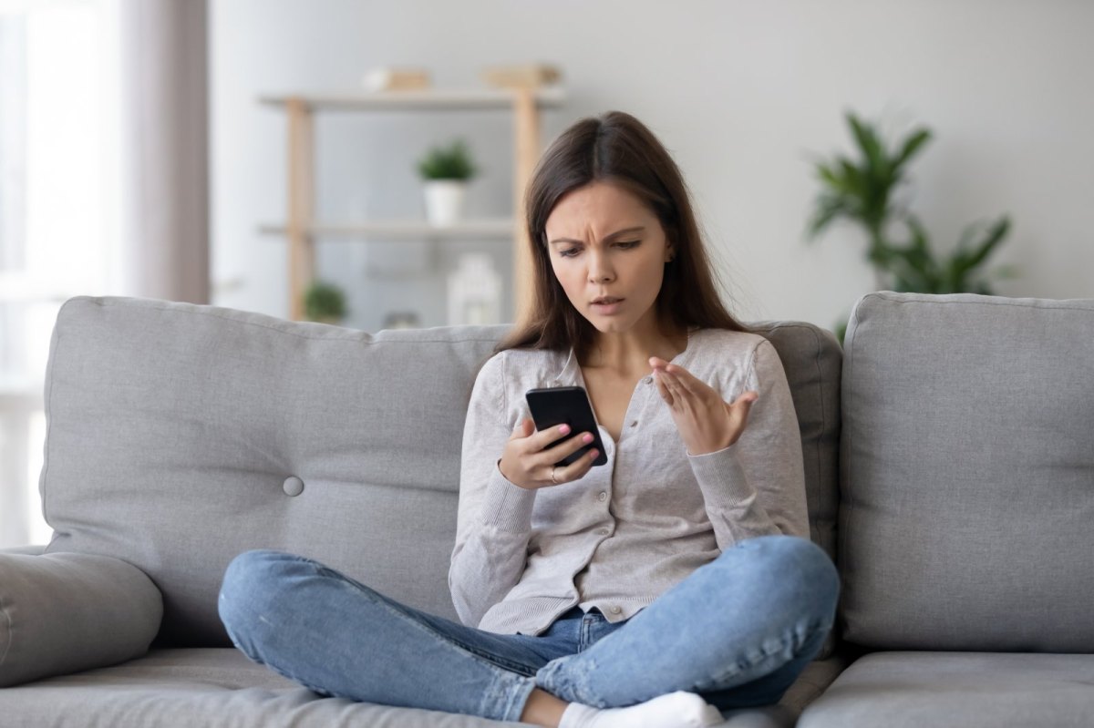 Frau sitzt mit ihrem Handy auf dem Sofa und schaut genervt auf das Gerät.