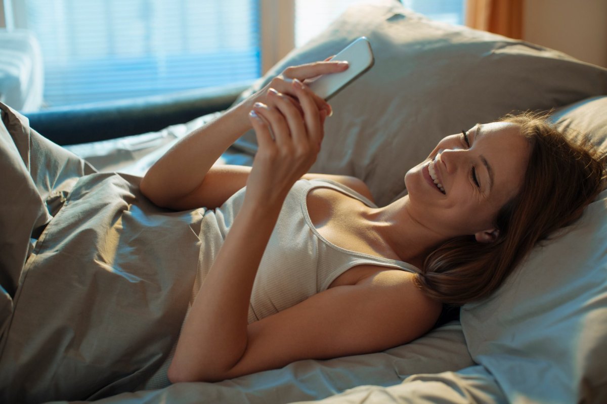 Frau liegt lachend im Bett und schaut auf ihr Handy.
