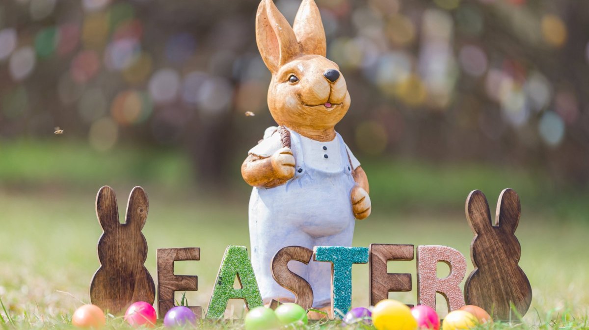 Osterhase steht hinter "EASTER"-Schild und Eiern.
