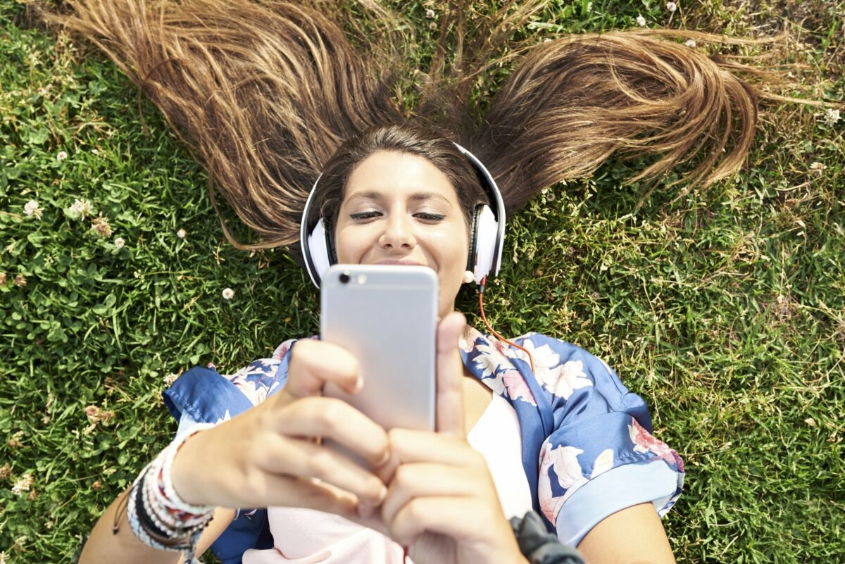 Eine Frau liegt mit Kopfhörern im Gras und schaut auf ihr Smartphone.