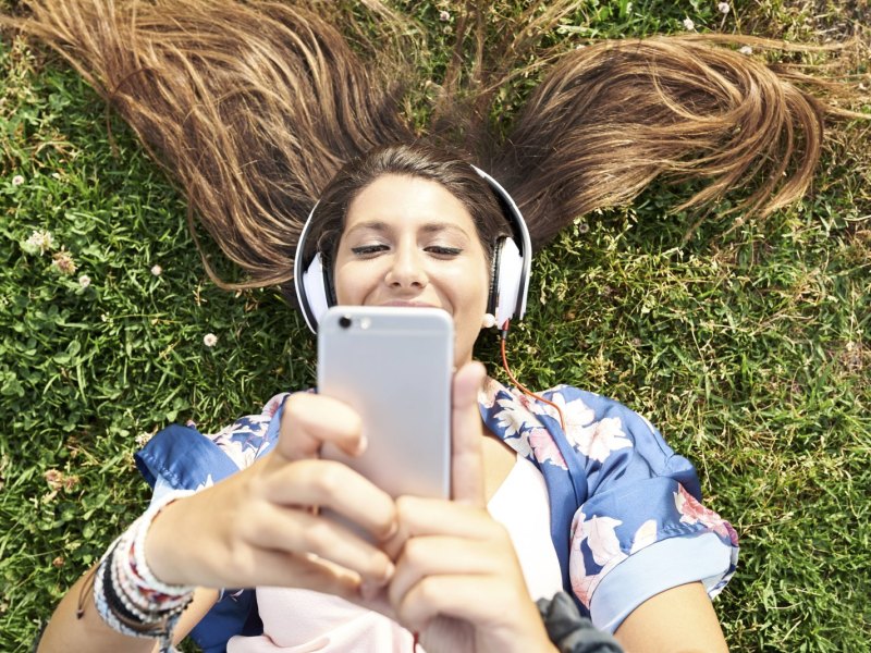 Eine Frau liegt mit Kopfhörern im Gras und schaut auf ihr Smartphone.