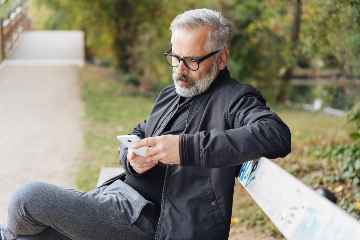 Mann sitzt mit iPhone auf Parkbank.