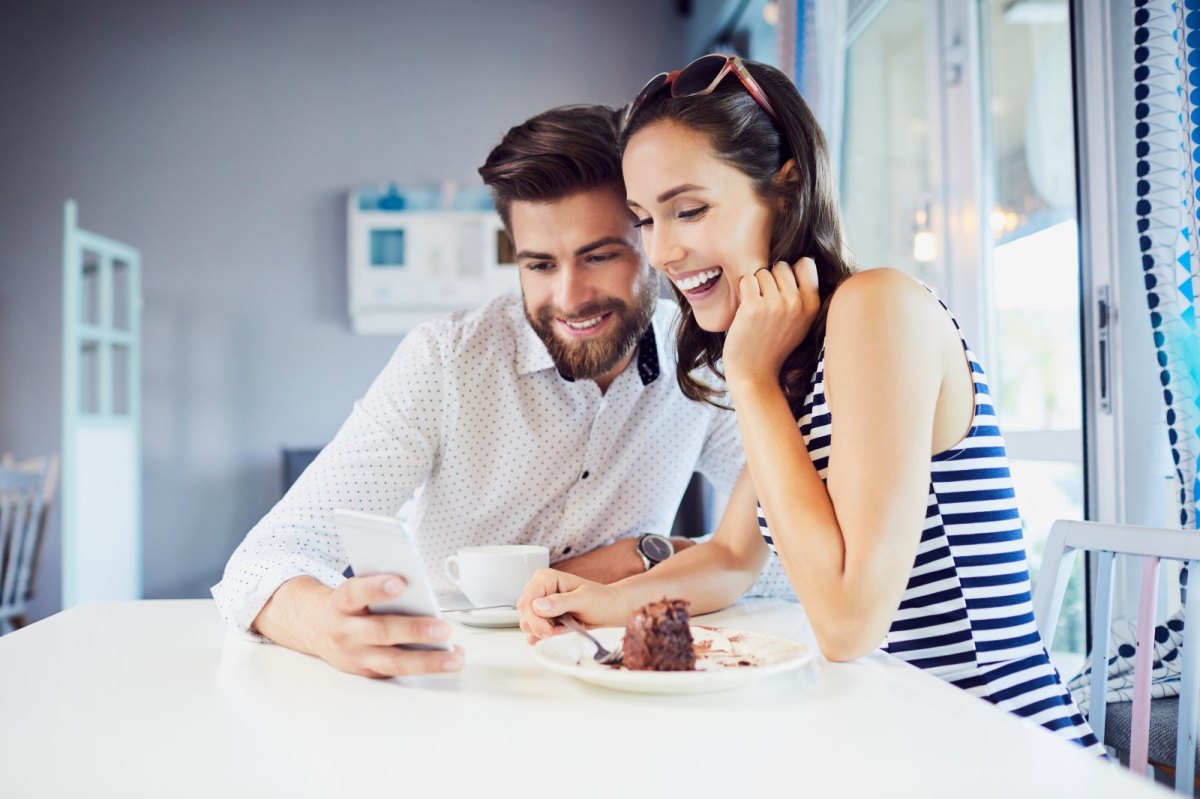 Ein Mann und eine Frau sitzen an einem Esstisch. Er zeigt ihr fröhlich etwas auf seinem Handy.
