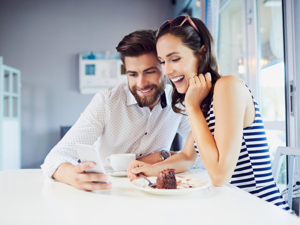 Ein Mann und eine Frau sitzen an einem Esstisch. Er zeigt ihr fröhlich etwas auf seinem Handy.