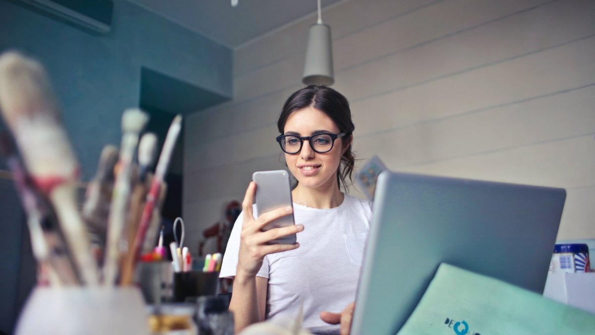 Eine Frau mit Brille schaut auf ihr Smartphone und sitzt an einem Schreibtisch.