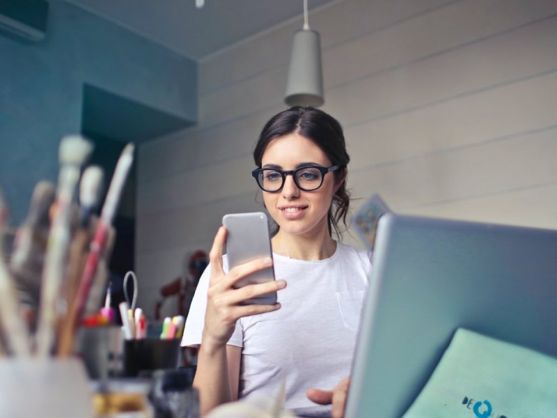 Eine Frau mit Brille schaut auf ihr Smartphone und sitzt an einem Schreibtisch.