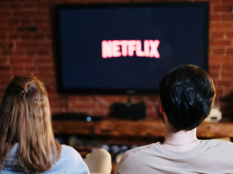 Zwei Menschen sitzen auf einem Sofa vor einem Fernseher und schauen Netflix.