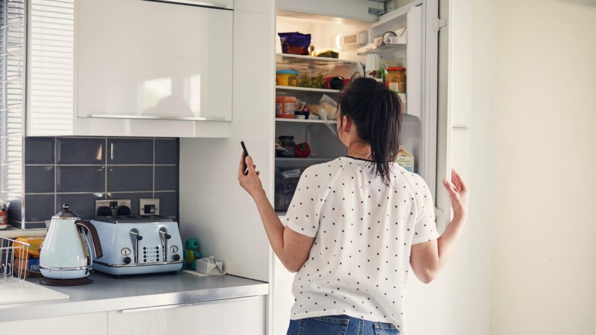 Frau mit Handy schaut in den Kühlschrank.