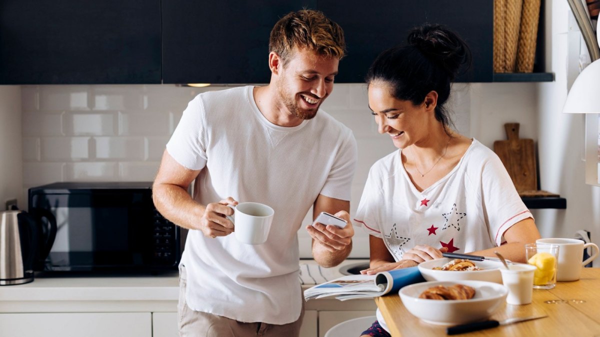 Ein Mann und eine Frau sind beim Frühstück in der Küche und schauen auf ein Smartphone. Sie lachen beide.