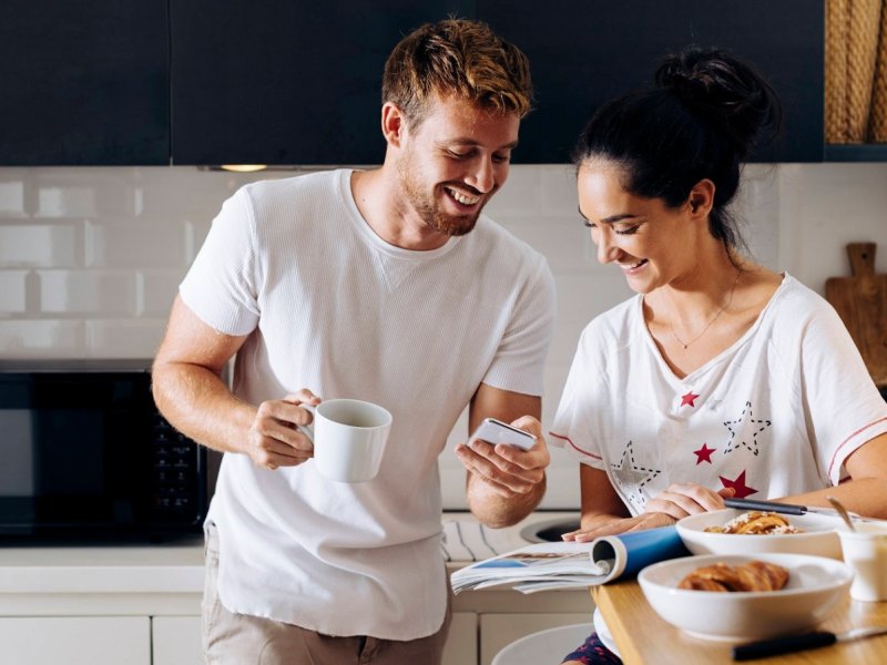 Ein Mann und eine Frau sind beim Frühstück in der Küche und schauen auf ein Smartphone. Sie lachen beide.