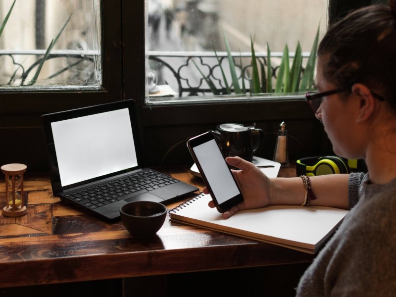 Frau sitzt vor Laptop mit Handy in der Hand