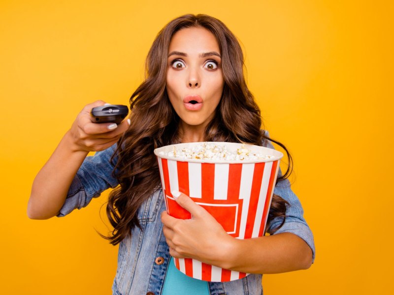 Frau mit Popcorn in der Hand drückt mit erstauntem Gesichtsausdruck auf eine Fernbedienung.