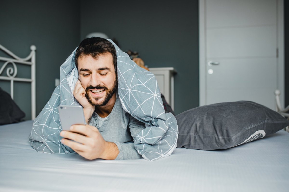 Mann liegt mit der Decke über den Schultern auf dem Bett und schaut fröhlich auf sein Handy.