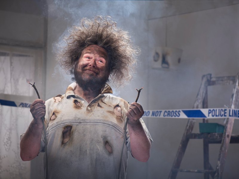 Mann mit abstehenden Haaren und rußbefleckt hält zwei verkohlte Kabel in der Hand.