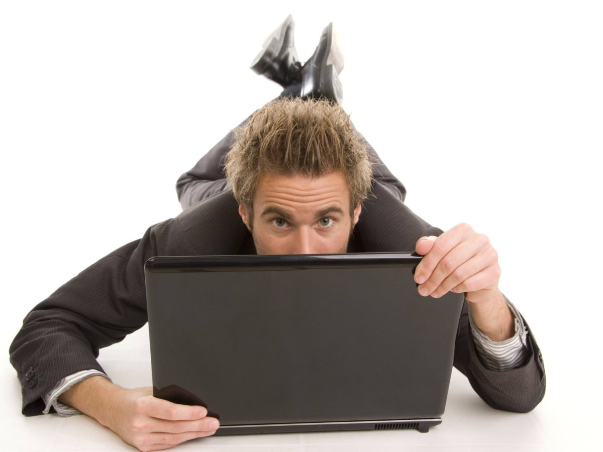 Mann im Anzug auf dem Bauch liegend versteckt sein halbes Gesicht hinter einem Laptop
