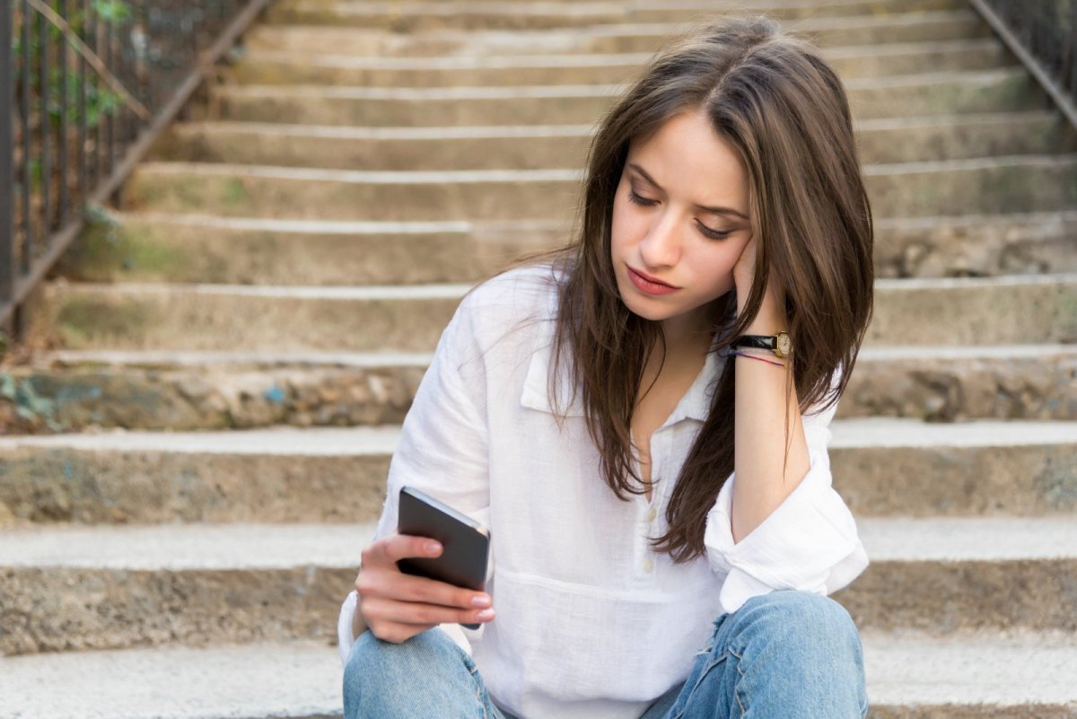 Frau sitzt auf einer Treppe und blickt traurig auf ein Handy.