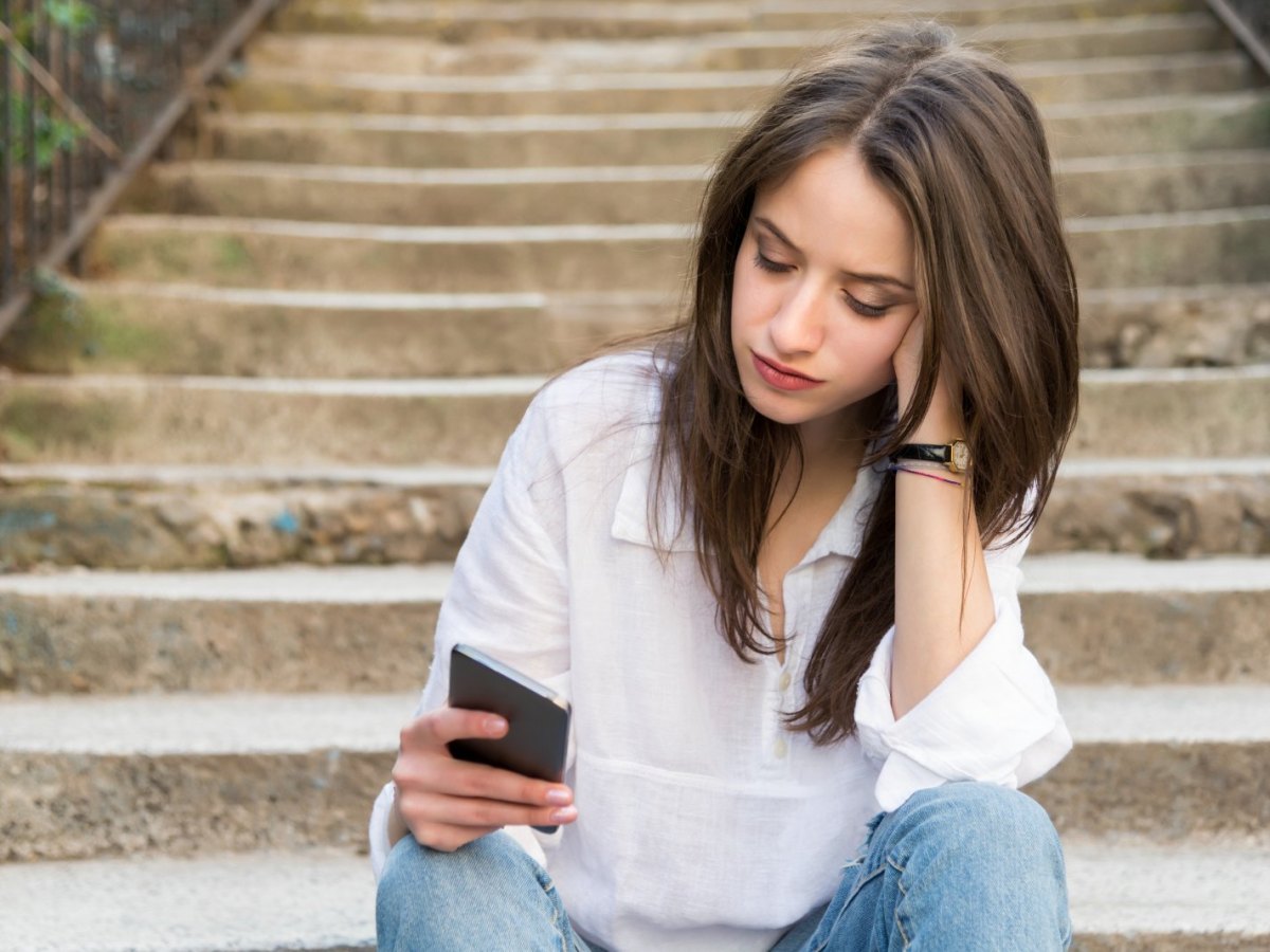 Frau sitzt auf einer Treppe und blickt traurig auf ein Handy.