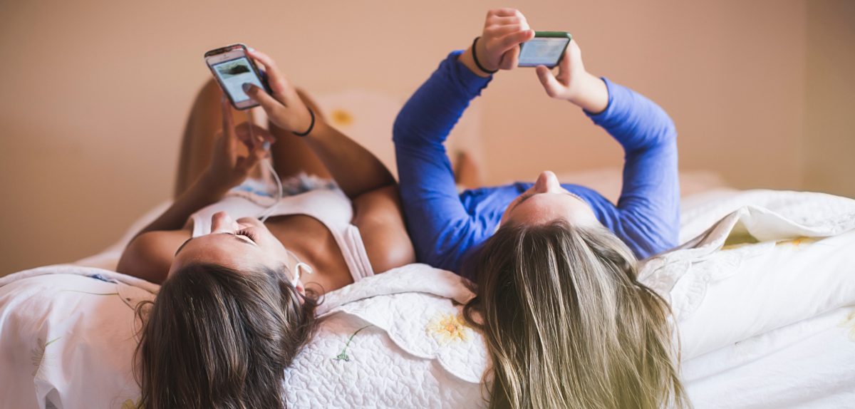 Zwei junge Frauen liegen rücklings auf dem Bett und schauen in ihre Samsung Galaxy S21 FE 5G Smartphones