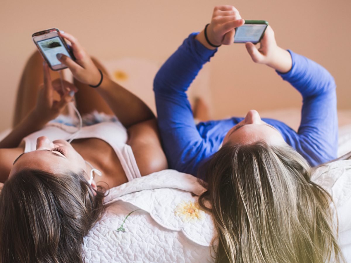 Zwei junge Frauen liegen rücklings auf dem Bett und schauen in ihre Samsung Galaxy S21 FE 5G Smartphones