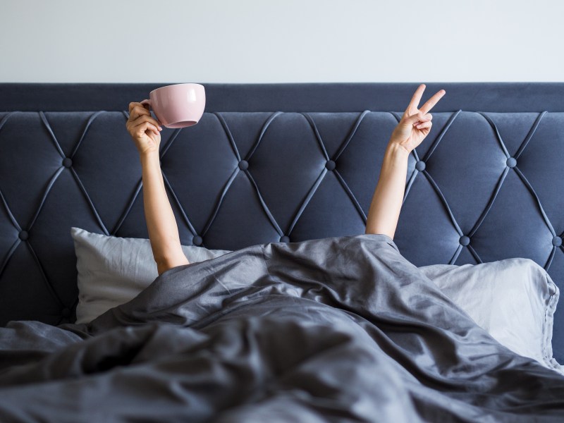 Frau im Bett hält Kaffee hoch