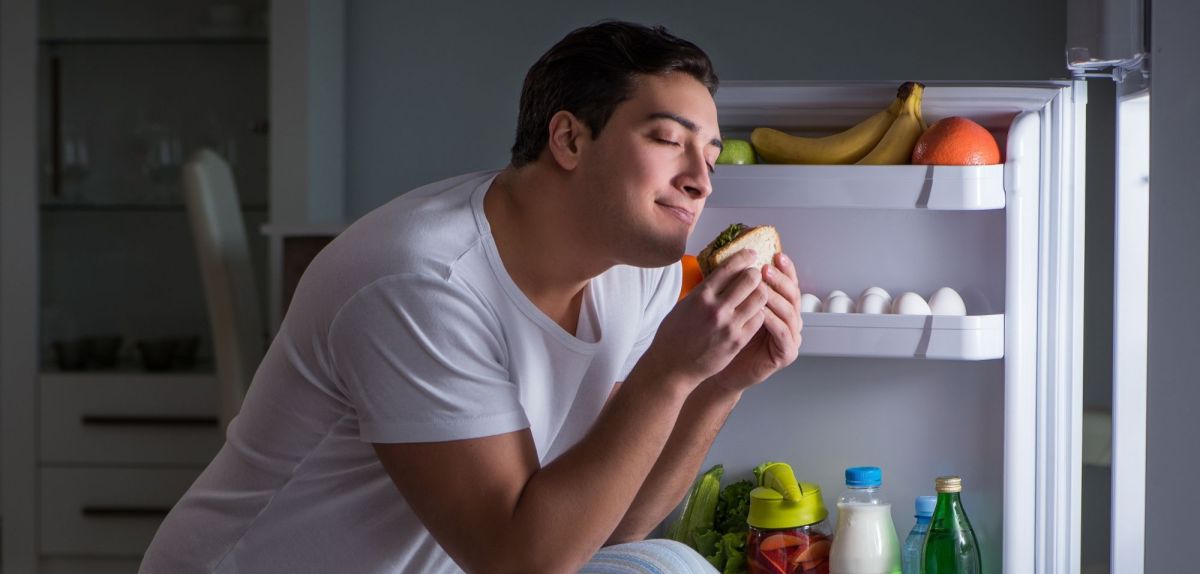 Mann isst Sandwich vor dem Kühlschrank.