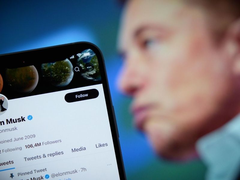 Bild von einem Handy mit Elon Musks Twitter-Profil, im HIntergrund Elon Musk.