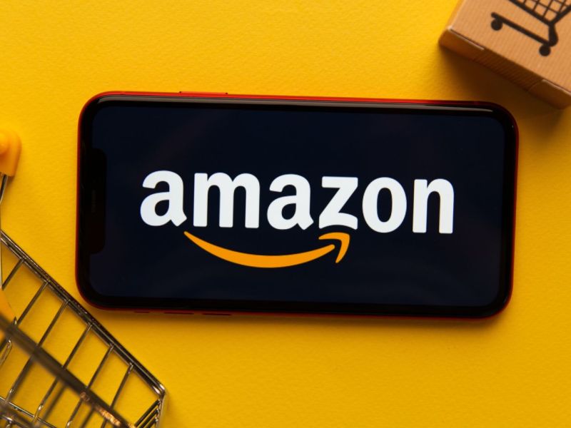 Amazon-Logo auf einem Handy zwischen Miniatur-Einkaufswagen und -Paket.