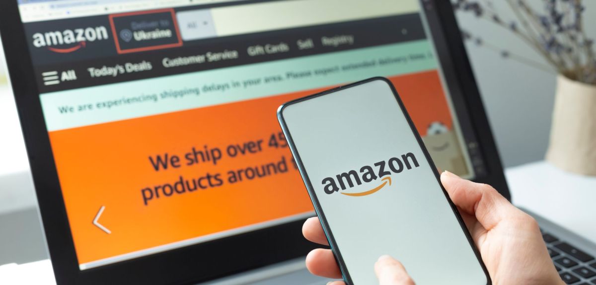 Person hält Handy mit Amazon-Logo auf dem Display in der Hand, dahinter ein Laptop-Bildschirm mit geöffneter Amazon-Webseite.