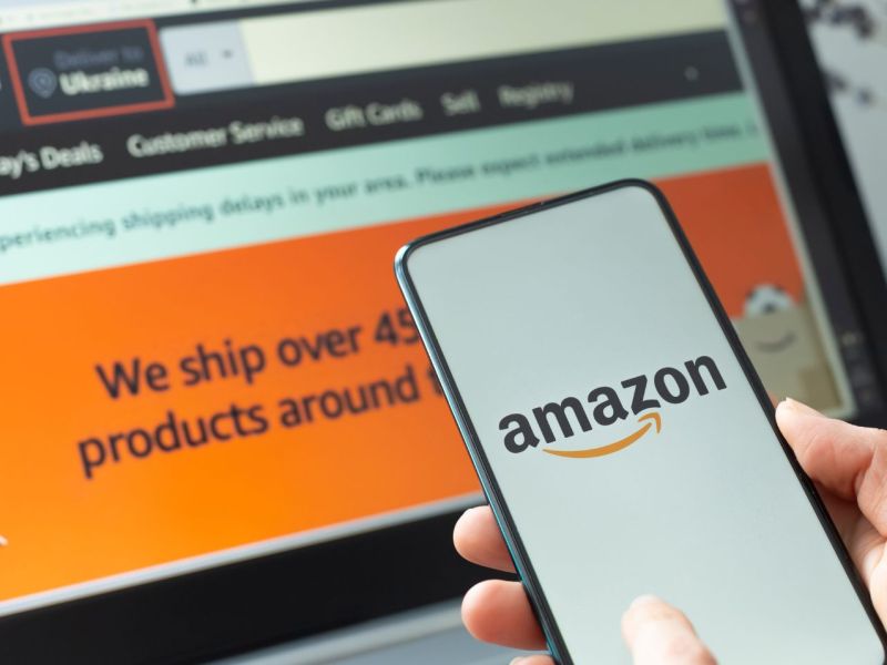 Person hält Handy mit Amazon-Logo auf dem Display in der Hand, dahinter ein Laptop-Bildschirm mit geöffneter Amazon-Webseite.