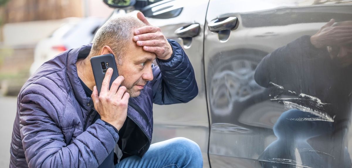 Mann telefoniert während er schockiert auf einen Kratzer an einem Auto schaut.