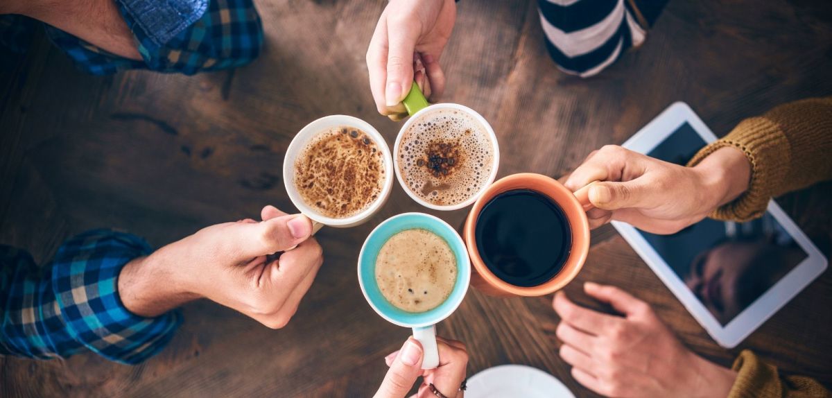 Mehrere Personen halten ihre Tassen mit Kaffee in die Mitte.