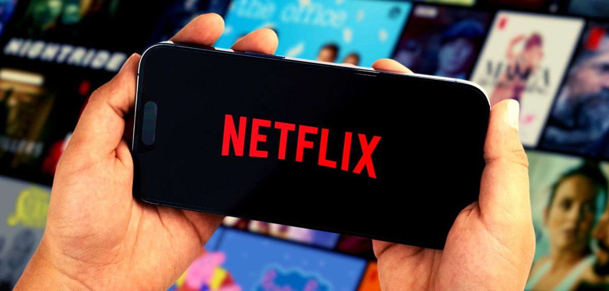 Person hält ein Handy mit dem Netflix-Logo in Händen. Dahinter sind Streaming-Kacheln zu sehen.