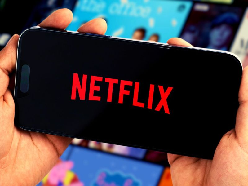 Person hält ein Handy mit dem Netflix-Logo in Händen. Dahinter sind Streaming-Kacheln zu sehen.