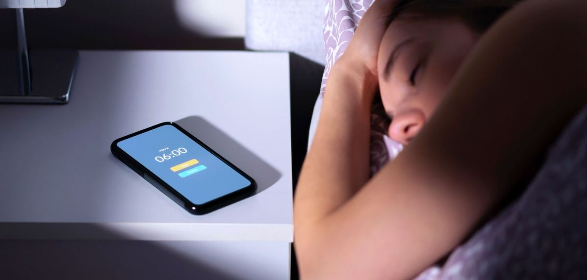Frau liegt im Bett und überhört den Wecker ihres daneben liegenden Handys.