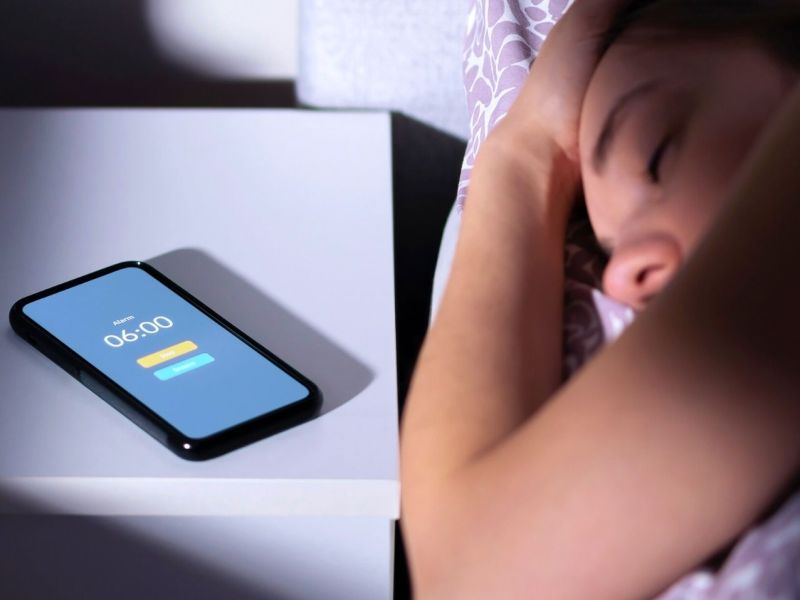 Frau liegt im Bett und überhört den Wecker ihres daneben liegenden Handys.