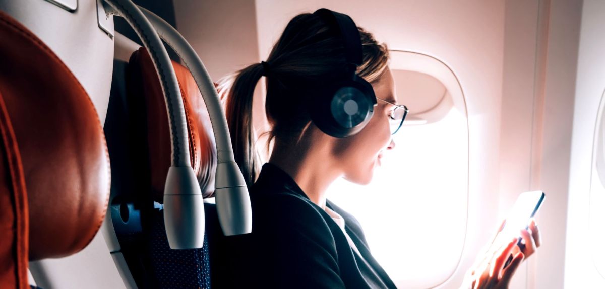 Frau sitzt mit aufgesetzten Kopfhörern im Flugzeug und schaut auf ihr Handy.