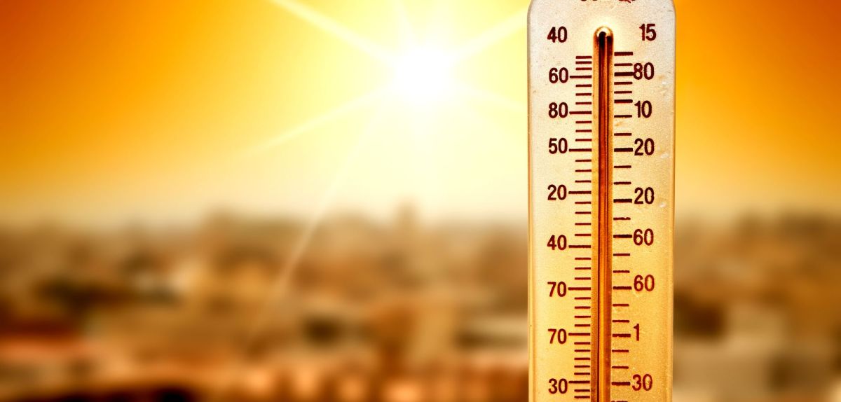 Ein Thermometer zeigt hohe Temperaturen an, im Hintergrund scheint die Sonne auf eine Stadt.