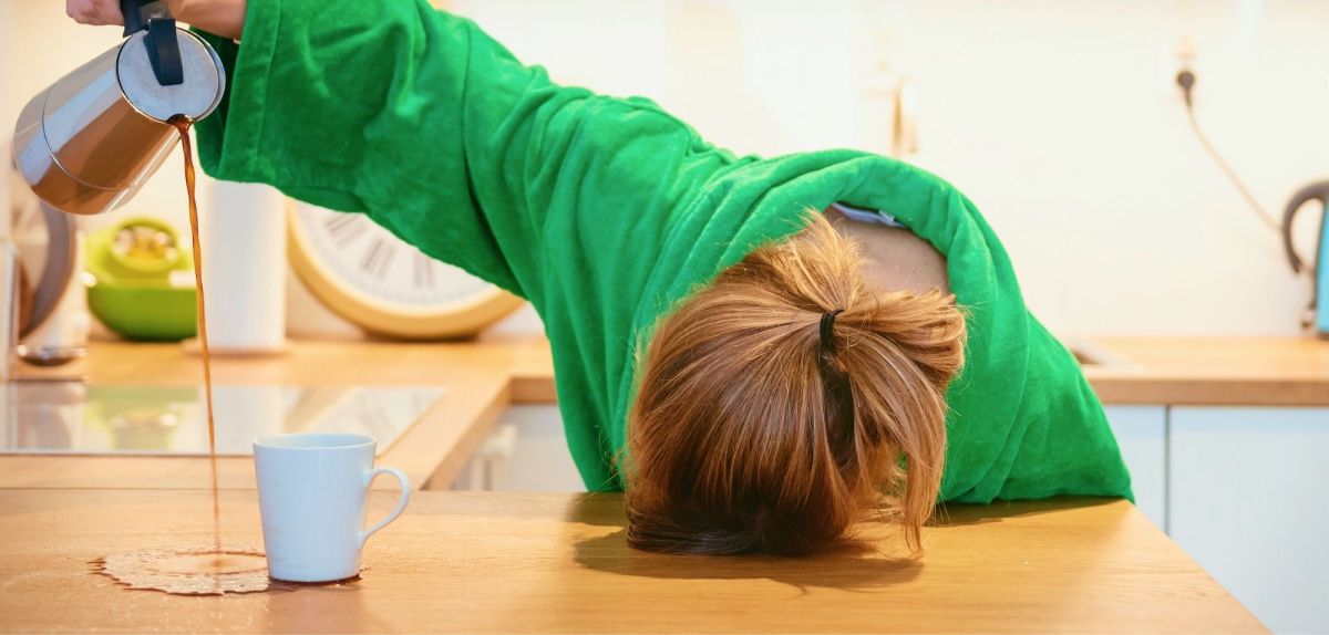 Übermüdete Frau mit Kopf auf dem Tisch gießt Kaffee neben Tasse.