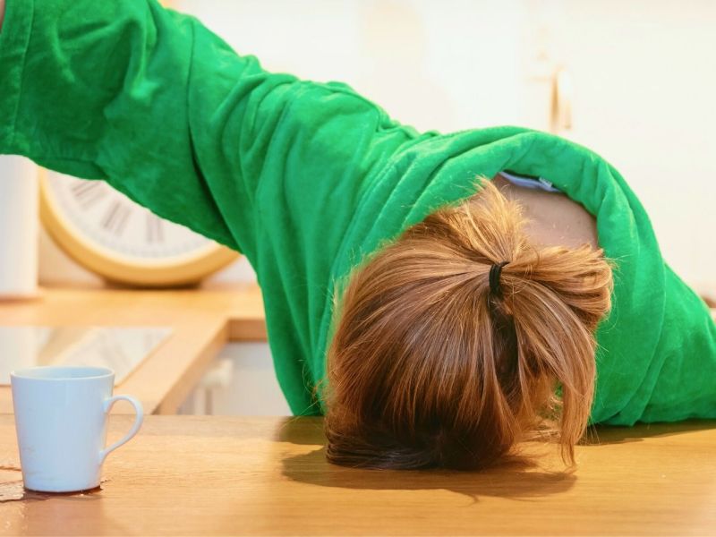 Übermüdete Frau mit Kopf auf dem Tisch gießt Kaffee neben Tasse.
