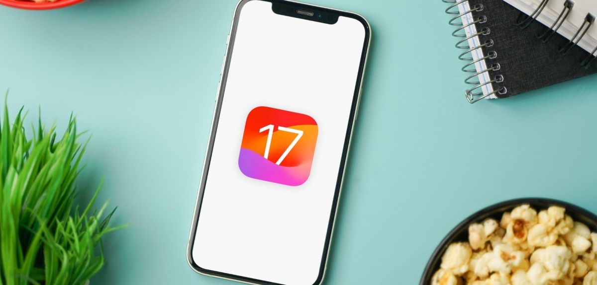 Handy mit iOS 17-Logo zwischen Popcorn-Schüssel, Notizheft und Pflanze.