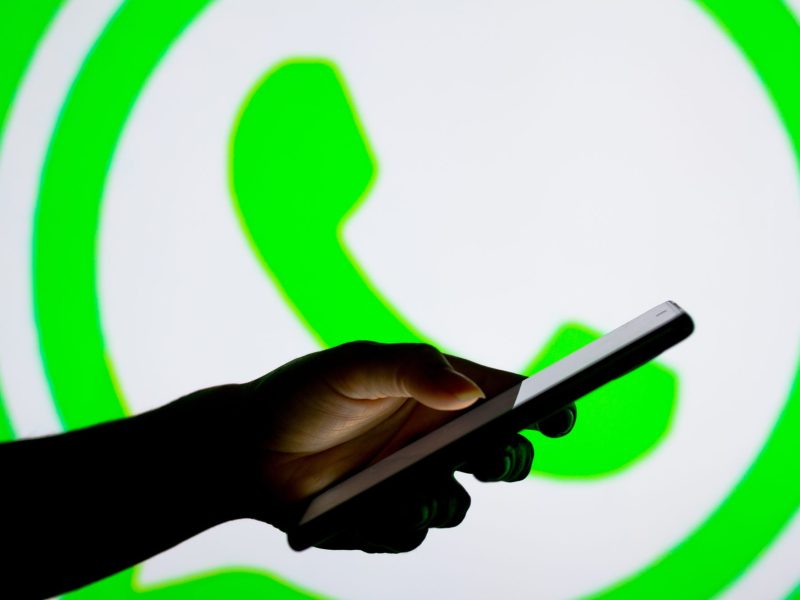 Eine Person hält ein Handy in der Hand, während im Hintergrund das WhatsApp Logo erscheint.