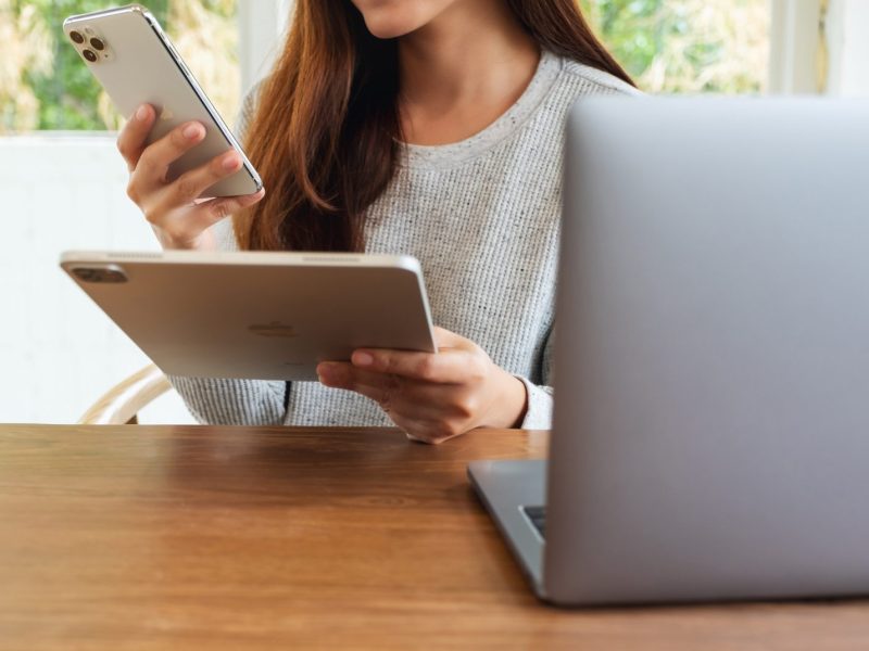 Frau hÃ¤lt iPhone und iPad von Apple, wÃ¤hrend sie vor einem Macbook sitzt.