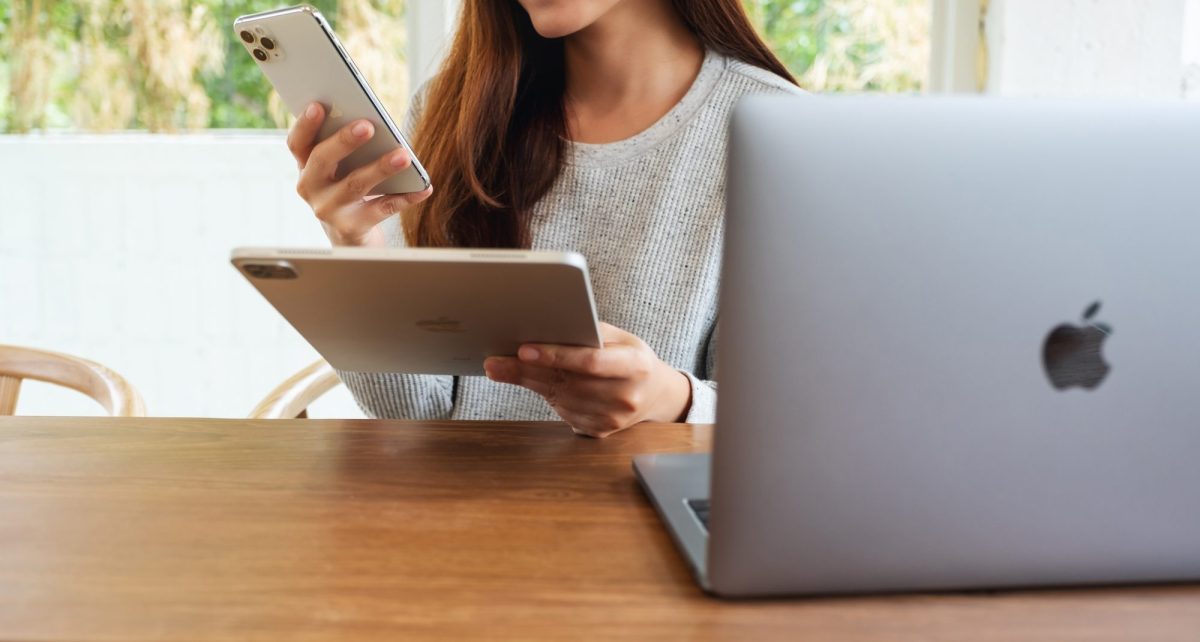 Frau hält iPhone und iPad von Apple, während sie vor einem Macbook sitzt.